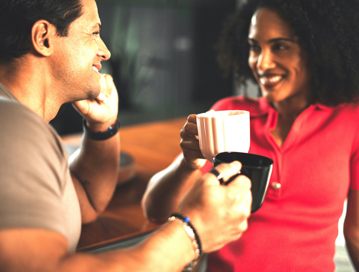 A smiling man and woman sitting close together indoors, each holding a mug. The woman wears a bright pink top, and the man wears a light beige shirt.