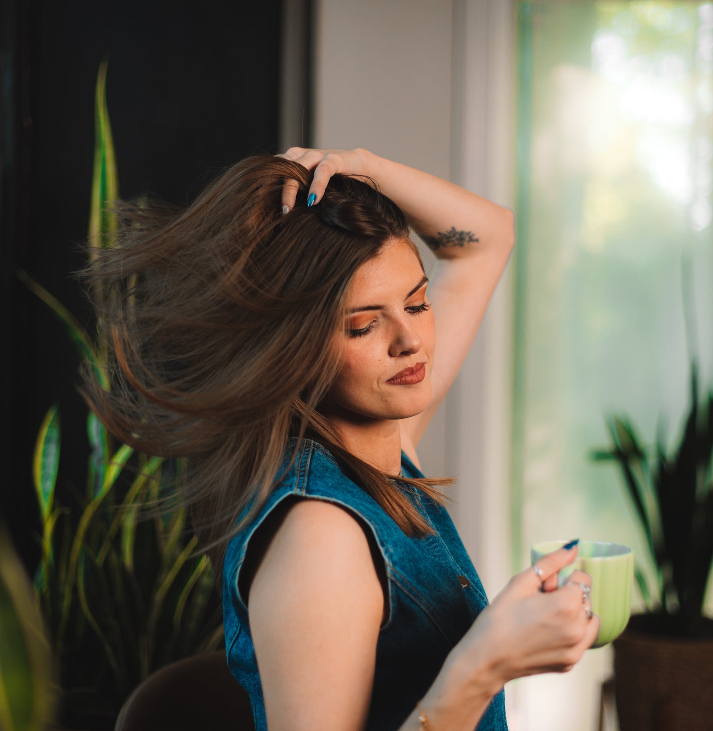 Woman holding a green mug with one hand and touching her hair with the other, standing indoors. She's drinking Dancing Ox Coffee